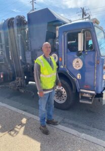 A man wearing a reflective safety vest standing next to a large blue city utility vehicle on a sunny day, appearing ready and professional