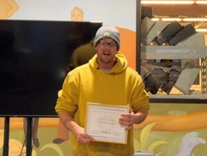 A man proudly holding up a certificate, wearing a yellow hoodie and beanie, standing in a colorful indoor setting during a recognition event.