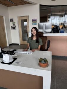 A smiling young woman with long dark hair, sitting at a reception desk in a modern office setting, with a computer, keyboard, and a small succulent plant nearby.