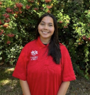 A young woman with long, dark hair smiling outdoors, wearing a red polo shirt with embroidered logos on the left side. She is standing in front of a lush green bush with red flowers, and the sunlight highlights her face and surroundings.