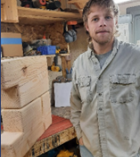 A young man with light brown hair and a beard standing in a woodworking workshop. He is wearing a beige long-sleeved work shirt and has a neutral expression. In the background, there are tools, wooden planks, and a workbench with large, neatly cut wooden beams.
