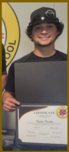 A young man with curly dark hair, smiling and holding a certificate in a black folder. He is wearing a black t-shirt and a black hard hat with a reflective emblem. The background features a circular logo on the wall, indicating a trade or training school.