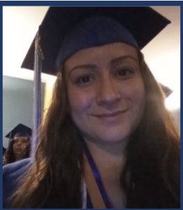 A woman with long brown hair, smiling in a blue graduation cap and gown. She is wearing honor cords around her neck. Another graduate in a blue cap and gown is visible in the background, slightly out of focus.