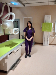 A young woman with long dark hair standing in a medical imaging room. She is wearing purple scrubs and black shoes, with her hands clasped in front of her. The room contains X-ray equipment, a large imaging table with a bright green surface, and a vertical imaging panel mounted on the wall.