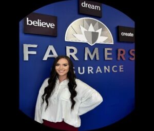 A smiling woman in front of a Farmers Insurance sign, wearing a white blouse and standing confidently. The backdrop includes motivational words: believe, dream, create.