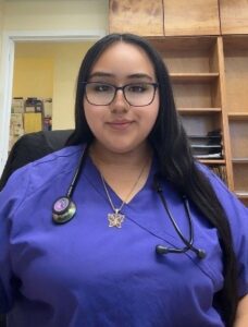 A young woman wearing glasses, a blue medical scrub top, and a stethoscope around her neck, seated indoors and smiling slightly