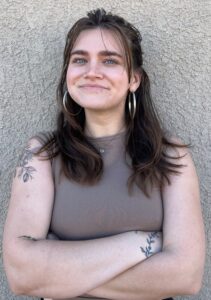 A confident young woman with long dark hair, large hoop earrings, and visible tattoos on her arms, standing with her arms crossed and smiling warmly against a textured wall