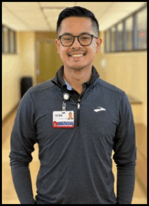 A smiling young man wearing glasses and a dark-colored quarter-zip jacket, standing in a brightly-lit indoor hallway with windows