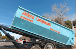 A large blue dumpster labeled 'Jackson Compaction' being loaded onto a truck outdoors, under a clear blue sky.