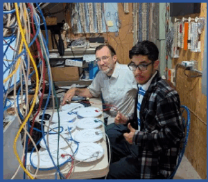 Two men sitting at a workspace with electronics equipment and multiple cables around them. They appear engaged in technical or network-related tasks