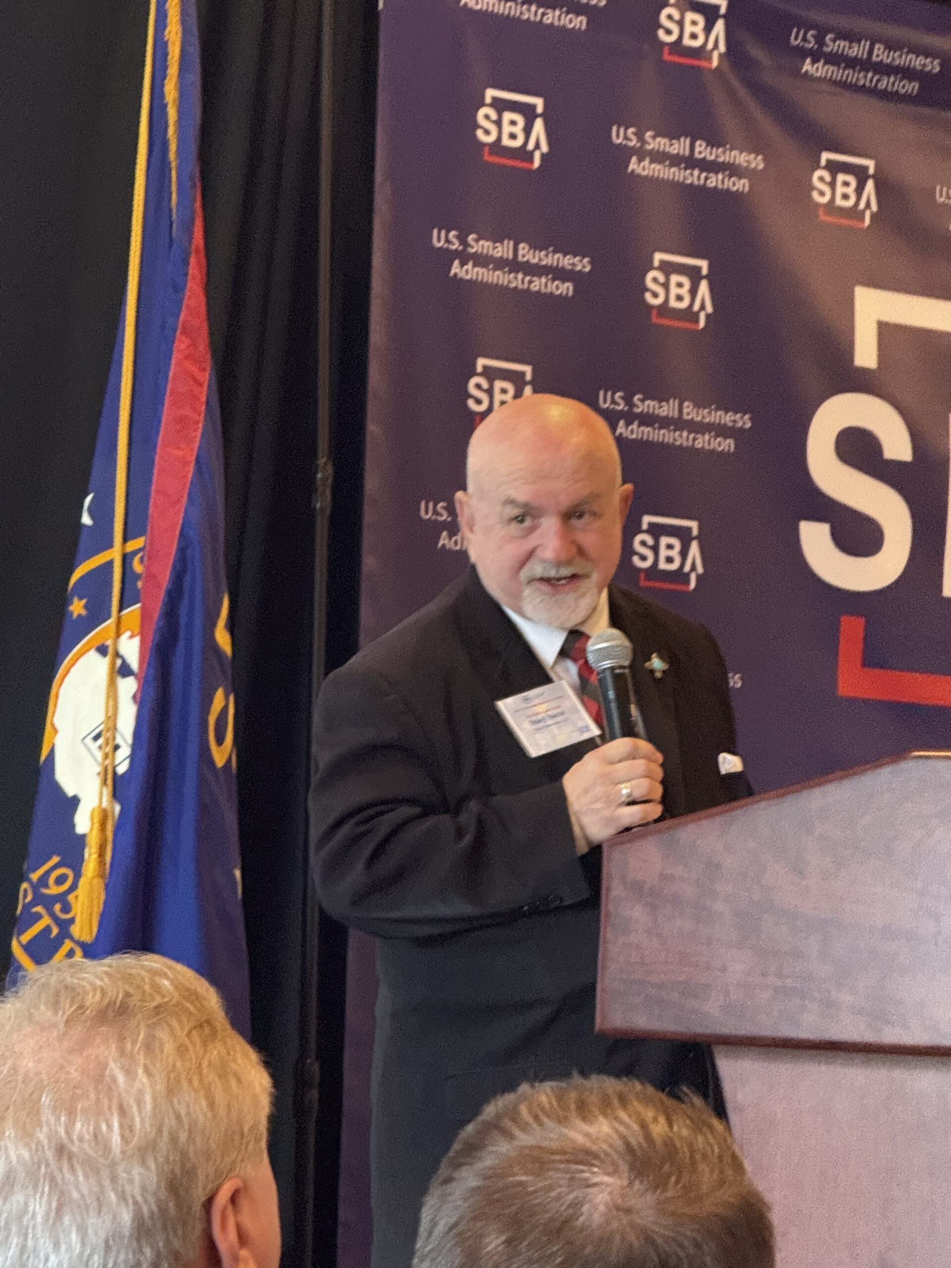 Man speaking at a podium during an SBA event, with U.S. Small Business Administration branding and U.S. flag in the background.