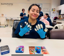 A young woman wearing blue medical scrubs and gloves on both hands, smiling at the camera in a classroom or training environment, with educational materials laid out on the table