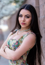 A confident young woman with long dark hair, wearing a floral dress and crossing her arms, posing outdoors beside a textured wall