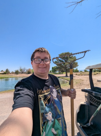 A smiling person wearing glasses and an anime-themed T-shirt, holding a garden rake outdoors, with a pond and greenery in the background