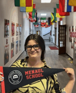 A smiling young woman wearing glasses and a patterned shirt, holding a pennant that reads 'Menaul School,' standing in a hallway decorated with international flags.