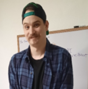 A young man wearing a backwards baseball cap and a blue plaid shirt over a black t-shirt, smiling slightly in front of a whiteboard.