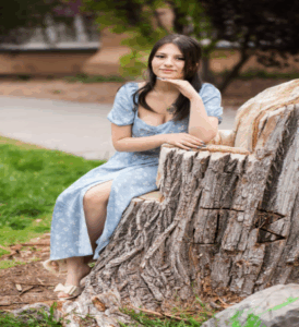 A young woman wearing a long, light-blue floral dress, sitting gracefully on a large tree stump in a park-like setting.