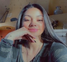 A smiling young woman with long dark hair, resting her chin on her hand, indoors with soft sunlight illuminating her face