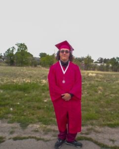 A graduate wearing a bright red cap and gown, proudly standing outdoors in an open field with trees in the background