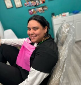 A smiling woman with dark hair, seated comfortably in a dental chair, wearing a pink dental bib in a brightly decorated dental office