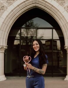 A smiling young woman with tattoos, wearing blue medical scrubs, holding a pink stethoscope shaped into a heart, standing in front of an elegant arched building entrance
