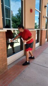 A person wearing a red shirt and shorts, cleaning windows outdoors on a sunny day, reflected clearly in the glass of the building.