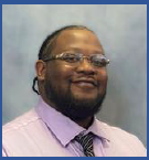 A man wearing glasses, a light purple shirt, and a striped tie, smiling in front of a neutral background.