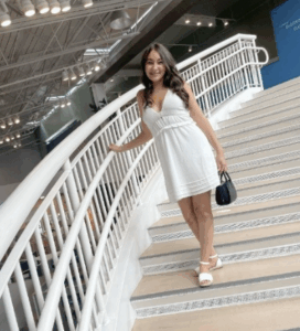 A smiling young woman wearing a white dress and heels, standing on a curved indoor staircase with modern lighting and railings