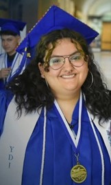 A smiling graduate wearing blue cap and gown with a white stole and medal, standing indoors with other graduates in the background.