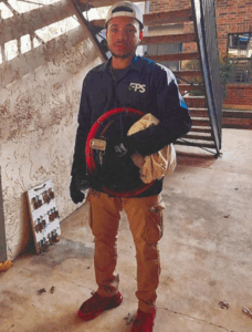 A man wearing a blue FPS jacket, tan cargo pants, and a backward cap, holding work equipment and standing near a staircase in an apartment complex.