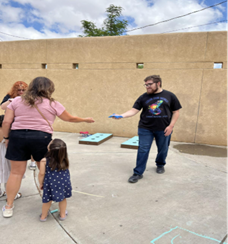 A man handing a beanbag to a woman during an outdoor activity while a small child stands beside her; cornhole boards are set up in the background.