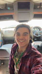 A young man with light brown hair, wearing a red, plaid unbuttoned shirt, smiling inside the cab of a semi truck.