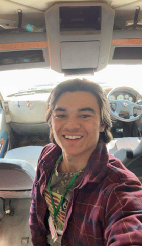A young man with light brown hair, wearing a red, plaid unbuttoned shirt, smiling inside the cab of a semi truck.