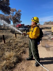 A young man, wearing fire fighter gear, holding a water hose, spraying water onto something out of the picture.