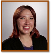 A woman with straight shoulder-length hair, wearing a black top and black blazer, smiling in front of a plain white background