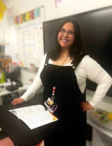A young female with long black hair, wearing glasses and black overalls and white long sleeve shirt, standing in a school classroom.