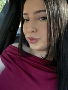 A woman with straight shoulder-length hair, wearing a dark pink top, smiling inside of a vehicle.