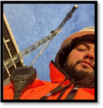 A man wearing a Florissant orange jacket and a worksite hard hat, standing in front of a large crane.