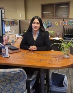A confident young woman with long dark hair, wearing a black dress, sitting at a desk with a computer off to the side