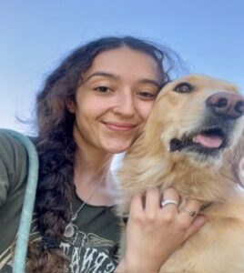 A confident young woman with long dark, braided hair, wearing a green shirt, petting a golden retriever dog, with the blue sky in the background