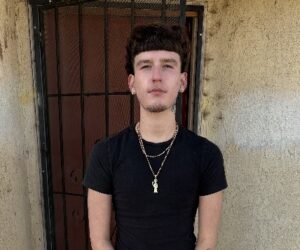 a confident young man with black hair, wearing a black shirt, standing outside of a building, in front of a large door