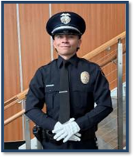 A young man with dark hair, wearing a full police uniform and white gloves, standing in front of a light brown staircase.
