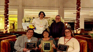 A young man with long brown hair, wearing a white button up shirt, standing in a resturant with four other individuals, holding plaques, wihile sitting in a booth.