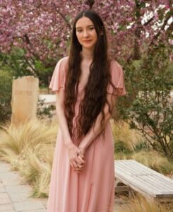 A confident young woman with long dark hair, wearing a pink dress dress and crossing her arms, posing outdoors beside a park bench