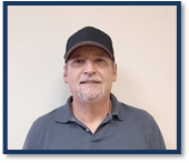 A man with a grey and white goatee standing in front of a white wall wearing a black baseball cap and a grey collared shirt.