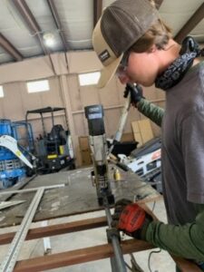 Person in a workshop using a power drill on a metal frame, wearing gloves and a cap, with tools and equipment in the background.