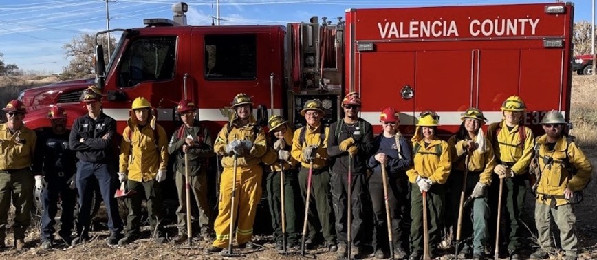 Group of firefighters in yellow and black gear standing with tools in front of a red Valencia County fire truck.